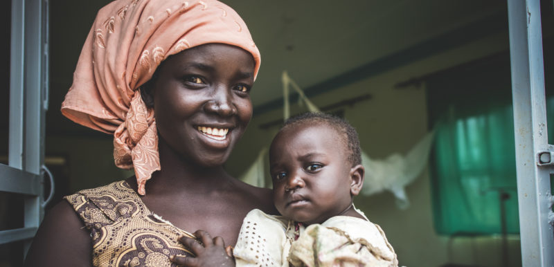 A Uganda refugee mother smiling while holding her baby in the hospital wing