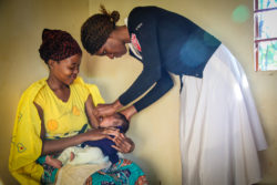 Young, smiling refugee woman holds her baby as a Medical Teams nurse examines him