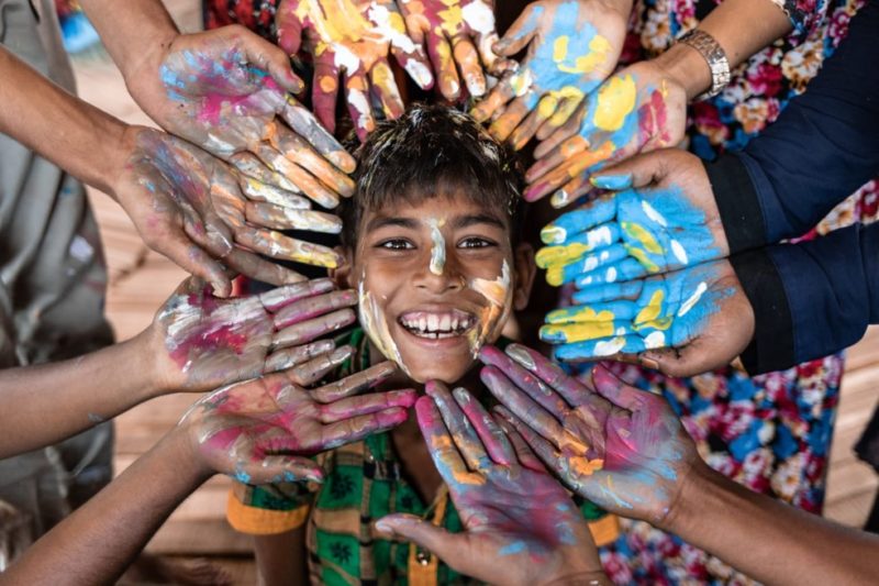 A Rohingya refugee boy covered in paint and smiling, with painted hands surrounding his face