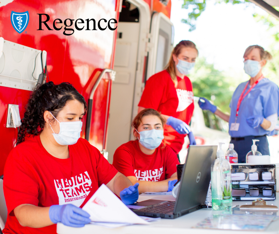 A group of women, wearing red shirts and face masks, gather around a table outside of a red mobile medical van and look at a laptop screen.
