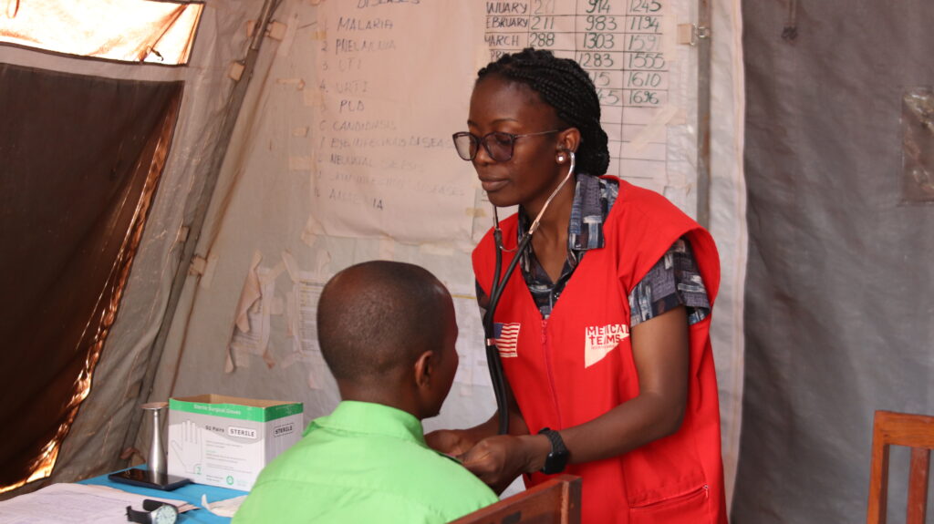 Dr. Eddah (standing) uses a stethoscope to check a patient (sitting).