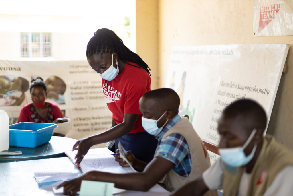 Racheal Kyalikoba stands next to two sitting men and looks at a book of records in a health facility.