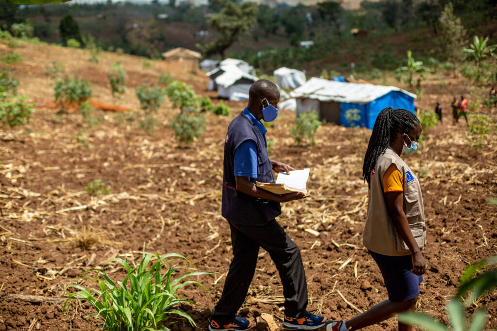 A man and woman walk through a field with tents in the background where refugees live.