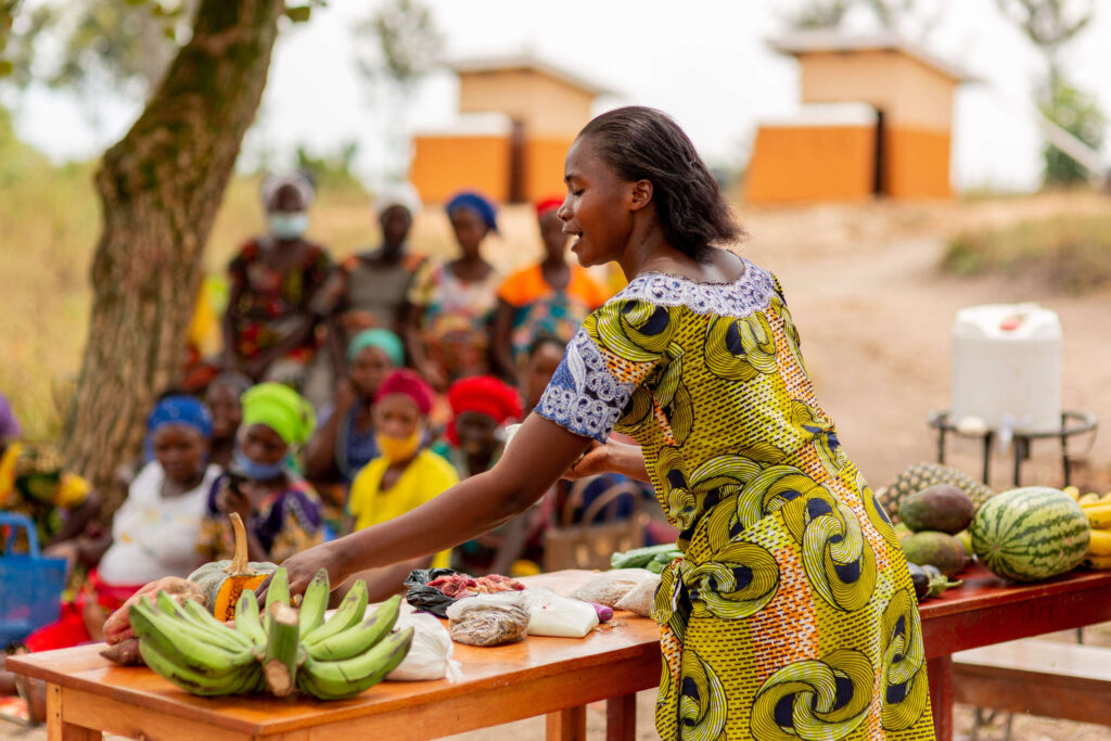 Florance stands in front of a group of women and shows fruits and vegetables.