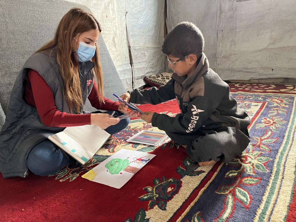 A young boy in Lebanon who struggled with his mental health draws with a young woman as they sit on a brightly patterned rug.
