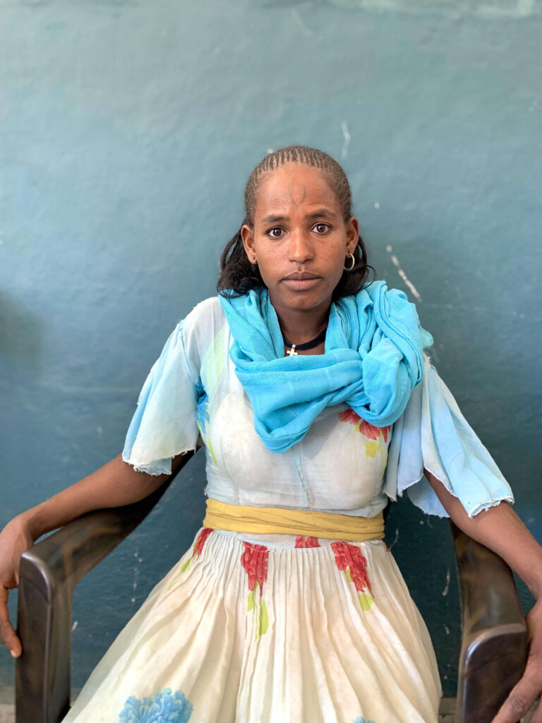 A woman in Ethiopia sits wearing a blue scarf in a chair against a blackboard.