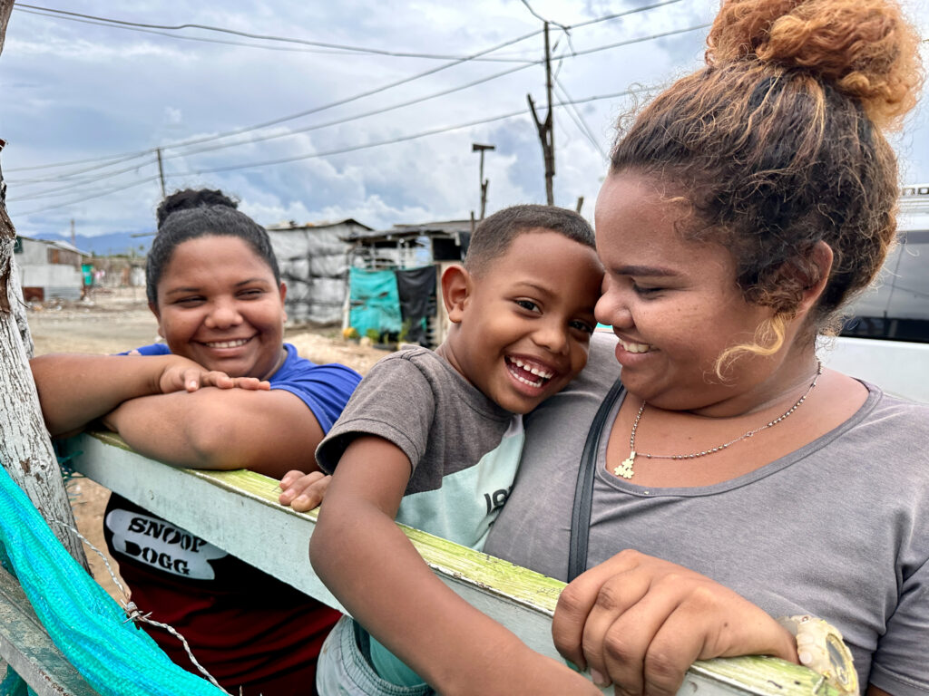 Two women smile at a little boy who's grinning at the camera. You can help people like them on Medical Teams Giving Day!