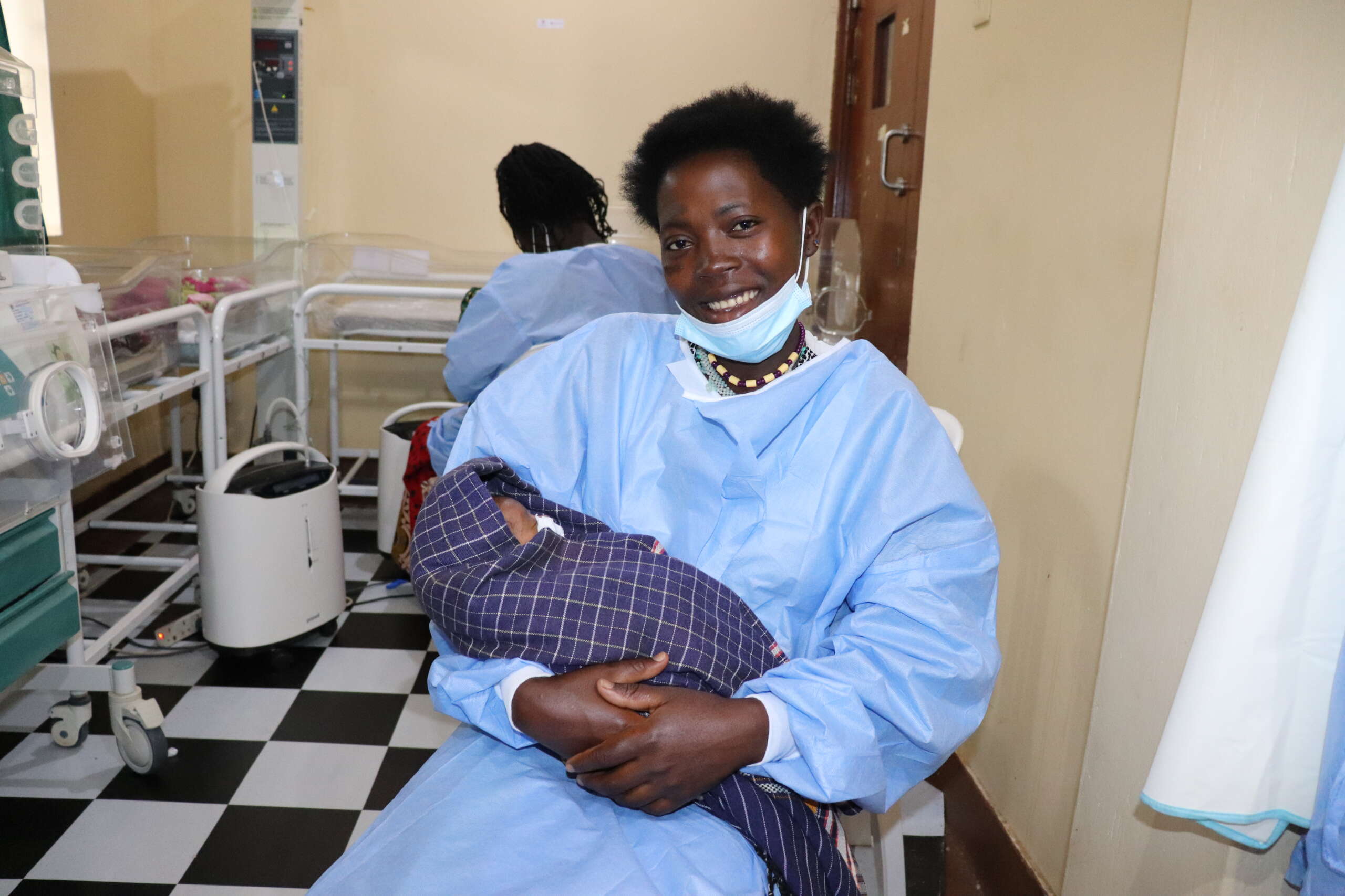 A mother in a surgery gown holds a baby in a NICU