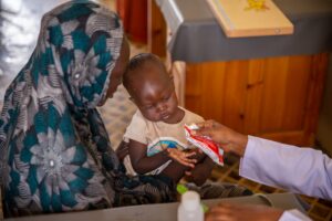 Ramadan sits on his mother's lap receiving nutrition-rich food from a health worker