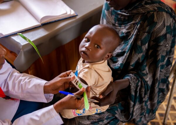 A health worker measures the arm circumference of a child named Ramadan using MUAC tape to check for malnutrition while he sits in the lap of his mother.